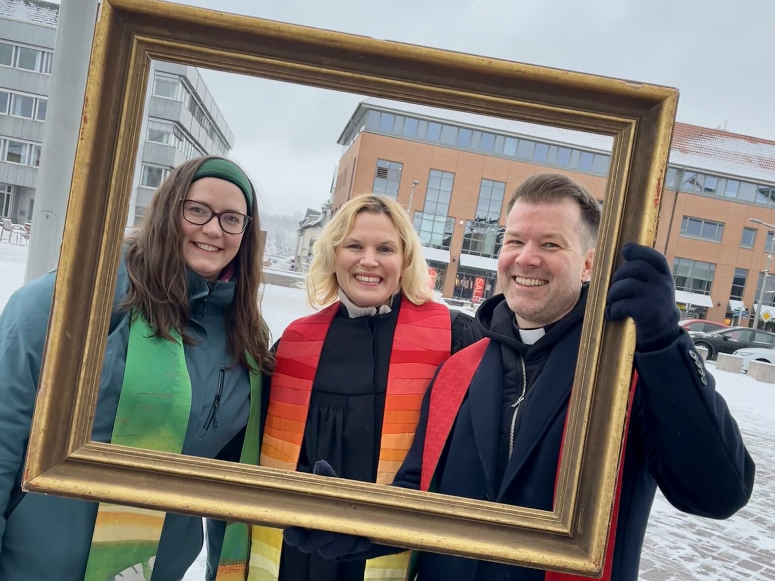 Drei fröhliche Menschen gucken durch einen großen Bilderrahmen in die Kamera.